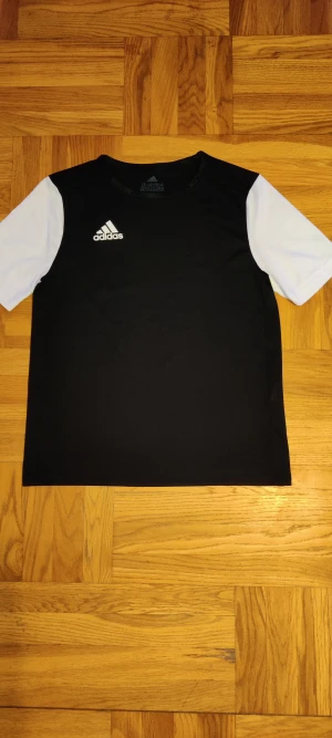 Svart och vit Adidas t-shirt - Snygg svart t-shirt från Adidas med vita ärmar och klassisk logga på bröstet. Tillverkad i ett lätt och sportigt polyestermaterial som andas. Perfekt för dig som gillar en stilren look och vill ha något bekvämt till träning eller vardag.