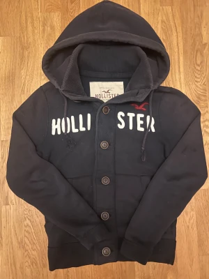Vintage Hollister cardigan / kofta med knappar - Marinblå hoodie från Hollister i storlek S med svart text på bröstet och Hollister-logga i vit. Tröjan har huva med snörning🔵|                                           Just dessa tröjor är väldigt eftertraktade!!                           Storlek: S längden är lite mindre i storlek   Skriv vid minsta fundering                                 Priset kan självklart diskuteras ! OBS!! Liten defekt vid inne i tröjan som man märker inte av när den är på! ( se bild ) och de tre nedersta knapparna är 