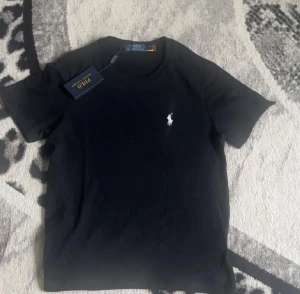 Svart t-shirt från Polo Ralph Lauren - Svart t-shirt från Polo Ralph Lauren med klassisk vit broderad logga på bröstet. Modellen är kortärmad och har en rund hals. Materialet är mjuk bomull som känns skönt mot huden. Perfekt för dig som gillar stilrena och tidlösa plagg. änvänt den max 20-25 gånger.