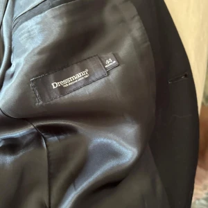 Svart kavaj från Dressmann, storlek 44 - Snygg svart kavaj från Dressmann i klassisk modell med två knappar framtill och fickor med lock. Fodret är slätt och har en diskret glans. Perfekt för dig som vill ha en stilren och tidlös look. Passar dig som gillar enkel och rak passform.