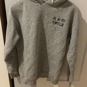 Grå hoodie från Gina Tricot - Grå hoodie från Gina Tricot med texten 'JE M'EN AMOUR' och en stjärna tryckt både fram och bak. 