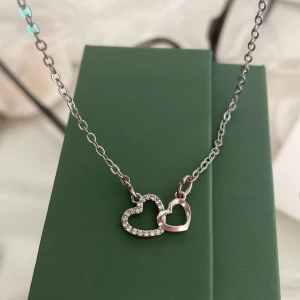 Halsband med dubbla hjärtan  - Snyggt halsband i silverfärg med två sammanlänkade hjärtan, där det ena hjärtat är dekorerat med små glittrande stenar. Perfekt accessoar för att lyfta din outfit med en söt och romantisk touch.