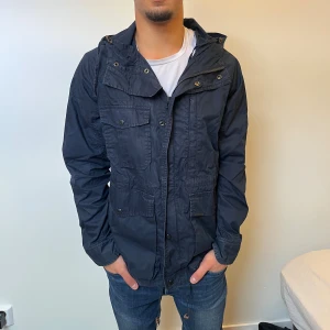 Mörkblå fieldjacka från Woolrich - Fieldjacka från Woolrich i mörkblått med huva, flera fickor med knappar och dragsko i midjan. Jackan har dragkedja och tryckknappar framtill, samt en klassisk streetstil. Perfekt för dig som vill ha en snygg och funktionell jacka till vardags. 