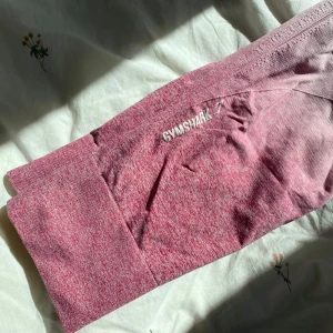 Rosa ombre leggings från Gymshark - Supersnygga seamless leggings från Gymshark i rosa ombre med hög midja. De är stretchiga och sitter tight, perfekta för gymmet eller annan träning. Materialet är mjukt och följsamt, och loggan syns diskret på låret. Ribbade detaljer längs benen för extra stil.