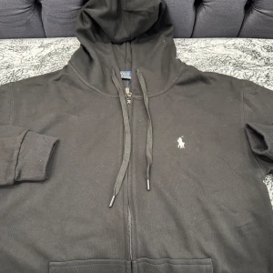 Svart hoodie från Polo Ralph Lauren - Svart hoodie från Polo Ralph Lauren med dragkedja, huva och snörning. Klassisk logga broderad på bröstet i vitt. Mjukt bomullsmaterial och ribbade muddar vid ärmslut och nederkant. Perfekt för en chill och stilren look.