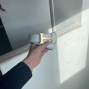 H bälte - Säljer ett stilrent vitt skärp från H med ett ikoniskt H-format spänne i guld. Bältet är tillverkat i äkta skinn och har en bred, klassisk form som verkligen sticker ut. Perfekt för dig som vill ha en lyxig accessoar med statement.