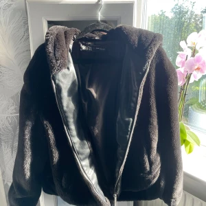 Svart teddyjacka med huva från Zara - Mysig svart teddyjacka från Zara i storlek XS. Jackan har en stor huva, dragkedja framtill och är tillverkad i mjukt pälsliknande material. Passformen är loose och jackan är perfekt för kyliga dagar. Snygg och enkel att matcha med olika outfits.