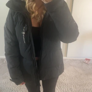 Svart pufferjacka från Sail Racing - Svart pufferjacka från Sail Racing med stor huva och dragkedja framtill. Jackan har logga på bröstet och ärmen, samt en clean och sportig look. Perfekt för kalla dagar och riktigt skön passform. Fodrad och med rejäla fickor. Den är i storlek L i barn storlek o passar mig perfekt som brukar ha xs,S i kläder. 