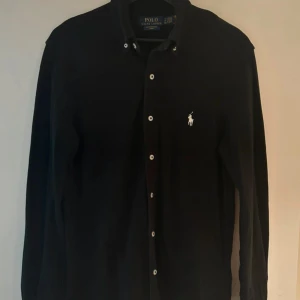 Svart långärmad skjorta Polo Ralph Lauren - Svart långärmad skjorta från Polo Ralph Lauren i tunn bomull. Klassisk krage, vita knappar och ikonisk broderad logga på bröstet. Perfekt för dig som gillar stilrena och tidlösa plagg med premiumkänsla.