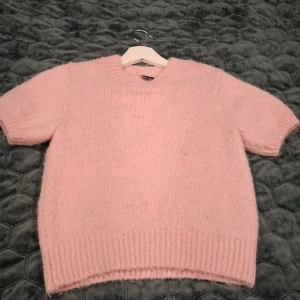 Ljusrosa stickad tröja Gina Tricot - Superfin ljusrosa stickad tröja från Gina Tricot med korta ärmar och rund hals. Tröjan har en mysig, fluffig känsla och ribbade muddar vid ärmslut och nederkant. Perfekt för lager-på-lager och en söt look.