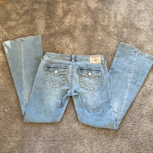 True Religion Joey ljusblå bootcut jeans - Ljusblå jeans från True Religion, modell Joey med låg midja och bootcut-ben. Klassiska detaljer med synliga sömmar, fem fickor och snygga knappar på bakfickorna. Tillverkade i mjukt denim-material för en avslappnad look. Jättefina i bra skick endast lite slitningar på övre lår och nere i kanten av byxan⭐️