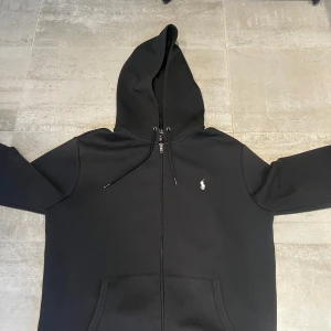 Svart hoodie från Polo Ralph Lauren - Svart hoodie från Polo Ralph Lauren med dragkedja och klassisk vit logga på bröstet. Tröjan har huva med snörning och känguruficka framtill. Perfekt för en chill och stilren look.