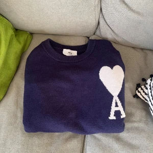 Mörkblå stickad tröja från Ami Paris - Mörkblå stickad tröja från Ami Paris med rund halsringning och lång ärm. På bröstet finns ett vitt hjärta och bokstaven A. Väldigt cozy tröja som är perfekt för vintern eller annan årstid.