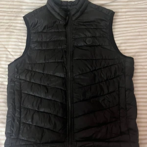 Svart dunväst från Jack & Jones - Svart quiltad dunväst från Jack & Jones med hög krage och dragkedja framtill. Västen har ett stilrent märke på bröstet och är tillverkad i syntetmaterial. Perfekt för lager-på-lager och enkel att matcha med olika outfits.