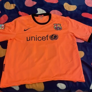 Barça Messi 10 orange fotbollströja Nike - Säljer en FC Barcelona fotbollströja i orange med svarta detaljer från Nike. Tröjan har korta ärmar, klubbmärke på bröstet, UNICEF-tryck fram och Messi 10 på ryggen. Materialet är lätt och andas, perfekt för match eller träning. Köpt tishan i Spanien.