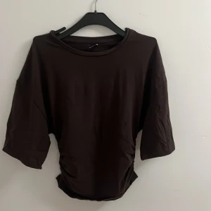 Brun topp med rynkade sidor - Mörkbrun topp med rund halsringning. Toppen har snygga rynkade detaljer i sidorna som ger en cool och modern look. Tillverkad i mjuk bomull som känns skön mot huden. Perfekt för dig som gillar en enkel men trendig stil.