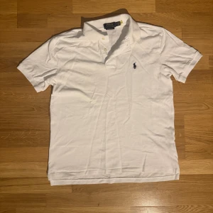 Vit pikétröja från Ralph Lauren - Klassisk vit pikétröja från Ralph Lauren med broderad marinblå logga på bröstet. Tröjan har korta ärmar, krage och knäppning med tre knappar. Modellen är Custom Slim Fit och tillverkad i mjuk bomull. Perfekt för en clean och stilren look.