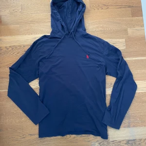 Mörkblå hoodie från Polo Ralph Lauren - Snygg mörkblå hoodie från Polo Ralph Lauren i mjuk bomull. Tröjan har huva med snörning, långa ärmar och den klassiska röda broderade loggan på bröstet. Perfekt för en chill och stilren look.