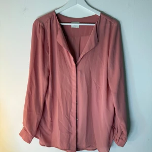Vila rosa blus  - Ljusrosa blus med långa ärmar och knäppning framtill. Blusen har en ledig passform och en enkel krage. Storlek M