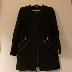 Svart kappa med dragkedjor  - Stilren svart kappa med raka linjer och flera snygga dragkedjor framtill och vid fickorna. Jackan har lång ärm och är perfekt för dig som gillar en minimalistisk look med coola detaljer.