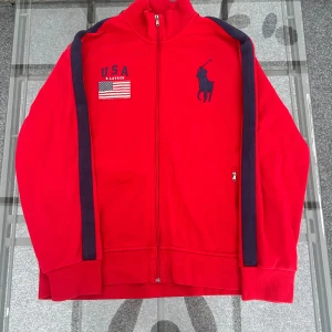 Polo Ralph Lauren röd ziptröja XL USA logga - Exklusiv Polo Ralph Lauren ziptröja i rött med blå detaljer och hög krage. Broderad USA-flagga + stor Polo-logga på ryggen gör den till en riktig statement-piece. Storlek XL, passar både oversized och true to size beroende på look. Perfekt för dig som gillar streetwear, sportig stil eller vintage Ralph Lauren. Äkta Ralph Lauren – superbra skick, inga skador eller fläckar.
