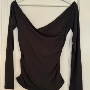 Svart omlottopp från Bik Bok - Svart långärmad topp med omlott-design och djup v-ringning. Toppens material är stretchigt och följsamt, vilket ger en snygg passform. Perfekt för dig som gillar en stilren och trendig look med lite extra edge.