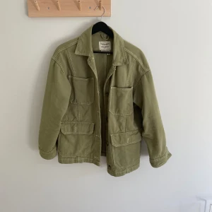 Grön jacka från Pull&Bear - Olivgrön jacka från Pull&Bear med fyra stora fickor framtill och knäppning med knappar. Jackan har klassisk krage, raka ärmar och synliga sömmar som ger en cool utility-vibe. Perfekt för lager-på-lager och chill stil.