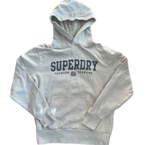 Vit hoodie från Superdry med tryck - Snygg vit hoodie från Superdry med stort mörkblått tryck på bröstet. Klassisk känguruficka framtill, ribbade muddar och huva. Perfekt för dig som gillar sportig och avslappnad stil.