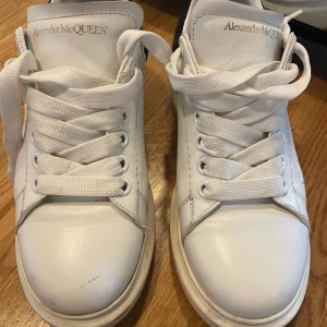 Alexander McQueen vita sneakers - Säljer ett par klassiska vita sneakers från Alexander McQueen med svart detalj på hälen. Skorna har tjock sula, rund tå och snörning. Tillverkade i skinn med stilren design och diskret logga på plösen. Perfekta för dig som gillar lyxiga och trendiga sneakers.