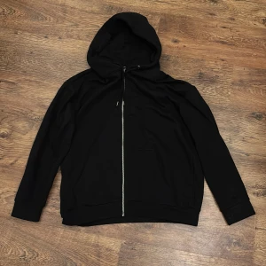 Svart  zip hoodie från Pier One - Svart hoodie från Pier One med hel dragkedja framtill och huva med snörning. Tröjan har långa ärmar, ribbade muddar och en klassisk känguruficka. Perfekt för en chill och stilren look.