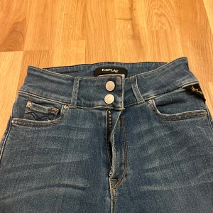 Blå Replay jeans bootcut - Snygga blå jeans från Replay med klassisk femficksmodell, dubbla silverknappar och dragkedja. Bootcut-modell med något utsvängda ben och hög midja. Tydliga Replay-detaljer på bakfickan och läderpatch i midjan. Perfekta för en avslappnad men trendig look.