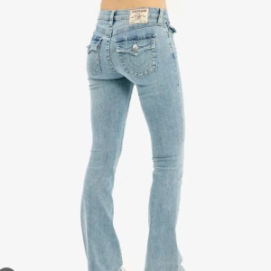 True Religion Becca Bootcut Jeans - Snygga ljusblå jeans från True Religion, modell Becca Mid Rise Bootcut. Jeansen har klassiska bakfickor med lock och dekorativa sömmar samt silverfärgade knappar. Tillverkade i jeansmaterial med en bootcut passform och normal midja.
