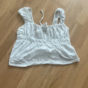 Vit linnetopp med volangärmar och knyt - Söt vit linnetopp med volangiga axelband och knytning framtill. Toppen har en lätt rynkad struktur och resår under bysten för en skön passform. Perfekt för varma dagar och ger en romantisk vibe till din outfit.