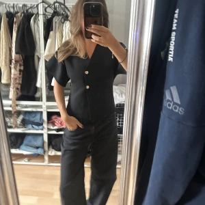 Svart blus med guldknappar - Säljer en svart blus med v-ringning och tre snygga guldknappar framtill. Blusen har korta, vida ärmar och en rak, något figurnära passform. Perfekt för dig som gillar stilrena och trendiga plagg med en twist.