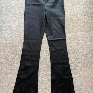 Svarta bootcut byxor i skinnimitation Mango - Snygga svarta bootcut byxor från Mango i skinnimitation. Byxorna har en smickrande passform med dragkedja i sidan och en mini slits längst ner vid kanden av fötterna. Tillverkade i ett mjukt syntetmaterial som ser ut som äkta skinn. Perfekta för dig som vill ha en edgy look.