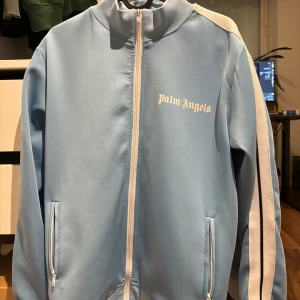 Palm Angels full-zip hoodie - Säljer en ljusblå vindjacka från Palm Angels med vita och svarta ränder längs ärmarna. Jackan har hel dragkedja framtill, ståkrage och två fickor med dragkedja. Materialet är mjukt och syntetiskt, perfekt för en chill streetwear-look.
