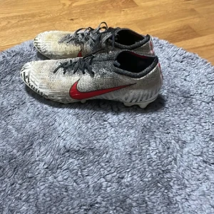 Nike fotbollsskor grå med röd swoosh - Säljer ett par Nike fotbollsskor i grått med svart och vit mönstring samt en röd swoosh på sidan. Skorna har snörning och platt sula med dobbar, perfekta för planen. Materialet är syntet och skon har en sportig, smal passform.