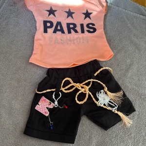 Persikofärgad topp med PARIS-tryck - Trendig persikofärgad topp med svart text 'PARIS FASHION' och tre stjärnor framtill, dekorerad med små glittriga stenar. Toppen har korta ärmar och en söt knytdetalj på axeln. Perfekt för dig som gillar statement-plagg och coola tryck.