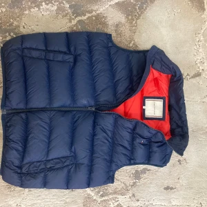 Mörkblå dunväst från Tommy Hilfiger - Snygg mörkblå dunväst från Tommy Hilfiger med röd insida och broderad logga på bröstet. Västen har två fickor framtill, quiltad design och dragkedja. Perfekt för lager-på-lager och streetstyle.