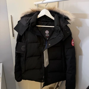 CanadaGoose Wyndham Svart - Helt ny CanadaGoose Wyndham jacka (svart), Jackan blev för liten på mig och då valde jag därför att istället sälja den här för ett rimligt pris. Jackan har aldrig använts och är exemplarisk bra för vinter varm och skön! med jackan så får du all tillbehör som justerbara päls-luvan som går att ta av, Canada Goose väska, prislapp mm. 