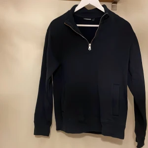 Svart half zip tröja från J.Lindeberg - Svart långärmad tröja från J.Lindeberg med half zip-dragkedja och diskret broderad logga på bröstet. Tröjan har en stilren design, hög krage och två fickor framtill. 
