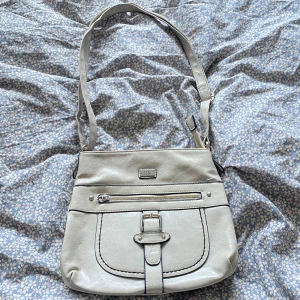 Ljusgrå axelväska från Feel - Snygg ljusgrå axelväska från Feel i syntetmaterial. Väskan har ett stort fack, dragkedja upptill och en dekorativ spänndetalj framtill. Perfekt storlek för vardagens essentials och justerbar axelrem för skön passform.