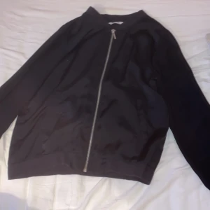 Svart bomberjacka i satin-look - Säljer en svart bomberjacka med glansig satin-finish och silverfärgad dragkedja framtill. Jackan har långa ärmar, ribbade muddar och diskreta fickor på sidorna. Perfekt för dig som vill ha en clean och stilren look.