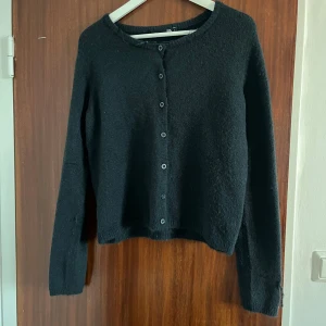 Svart kofta från 157 med knappar - Mysig svart kofta från 157 med rund halsringning och knäppning framtill. Koftan är stickad och har långa ärmar, perfekt för lager-på-lager under kyliga dagar. Enkel och stilren design som passar till det mesta.