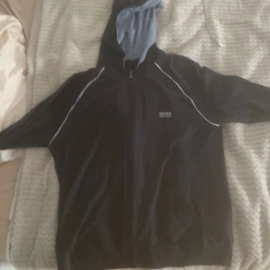 Svart hoodie från BOSS med dragkedja - Svart hoodie från BOSS med vit piping längs ärmarna och ljusblått foder i huvan. Tröjan har dragkedja framtill och BOSS-logga på bröstet. Perfekt för en chill och sportig stil.