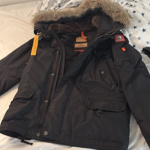 Svart Parajumpers vinterjacka med päls - Grym svart vinterjacka från Parajumpers med päls på huvan, flera fickor och snygga detaljer som gul rem och patch på ärmen. Slim fit-modell, perfekt för kalla dagar. Jackan har dragkedja och knappar framtill samt mjukt foder. Sist använt 2023. Använt 1 vinter