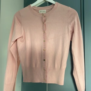 Ljusrosa kofta från Holly & White - Supersöt ljusrosa kofta från Holly & White med rund halsringning och knappar framtill. Långärmad och i mjuk bomullsblandning, perfekt för lager-på-lager. Enkel och stilren design som passar till allt.