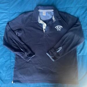 Mörkblå pikétröja Classic League - Mörkblå långärmad pikétröja från Classic League med vit knappslå och broderad logga på bröstet. Klassisk krage med randiga detaljer på insidan. Perfekt för en sportig och avslappnad stil.