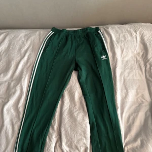 Gröna Adidas mjukisbyxor med vita ränder - Snygga gröna mjukisbyxor från Adidas med klassiska vita ränder längs sidorna och broderad logga på låret. Byxorna har resår i midjan och dragkedjefickor. Perfekta för chill eller träning, riktigt sköna och stilrena.