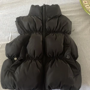 Svart dunväst från FB Sister XS - Svart puffig dunväst från FB Sister i storlek XS. Västen har hög krage, dragkedja framtill och två fickor med knappstängning. Snygg och trendig modell som ger en cool siluett. Perfekt att slänga över en hoodie eller tröja för extra värme.